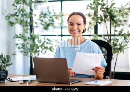 Erfolgreiche, intelligente Geschäftsfrau mit gemischter Rasse, finanzchefin, Analytikerin, sitzt an einem Schreibtisch in einem modernen Büro, hält Finanzdokumente in den Händen, blickt auf die Kamera und lächelt glücklich Stockfoto