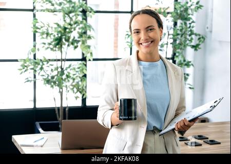 Foto einer positiven erfolgreichen selbstbewussten brasilianischen oder hispanischen lockigen Frau, makler, Top-Manager, Rekrutierer, hält ein Dokument in den Händen und eine Tasse, steht im Büro, schaut auf die Kamera, lächelnd Stockfoto