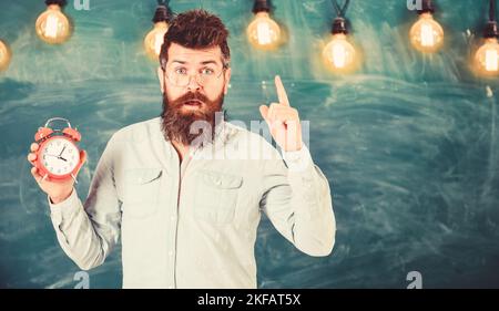 Der Lehrer in der Brille hält einen Wecker. Bärtiger Hipster hält die Uhr, Warnung mit Zeigefinger, Kopierraum. Disziplinkonzept. Mann mit Bart und Stockfoto