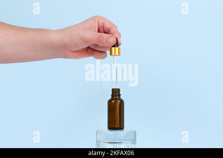 Männliche Hand hält Tropf mit Öl. Geöffnetes und geschlossenes Glas mit Naturkosmetikprodukt auf blauem Hintergrund. Ätherisches Öl für die Pflege der Haut. Hautpflege, Stockfoto