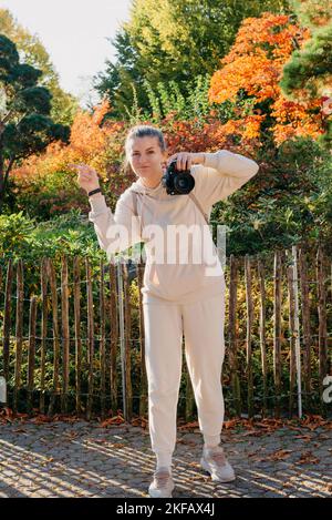 Schöne junge Frau macht Fotos mit einer professionellen Kamera im Herbstwald. Lächelndes Mädchen genießen Herbstwetter. Ruhe, Entspannung, Lifestyle Stockfoto