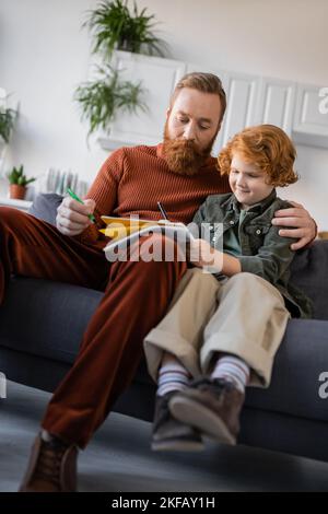 Rotschopf Junge schriftlich in Copybook in der Nähe bärtigen Vater auf der Couch zu Hause Stockfoto