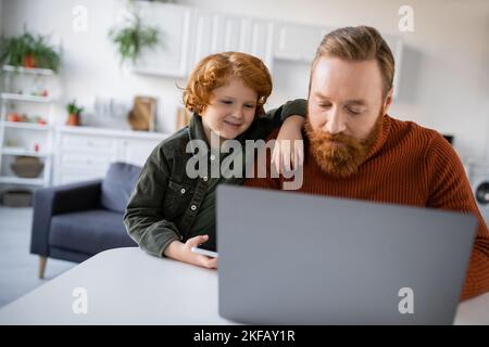 Rotschopf Kind lächelnd in der Nähe bärtiger Vater arbeitet auf verschwommenem Laptop zu Hause Stockfoto