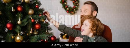 Rotschopf Junge aus Zunge und Dekoration Weihnachtsbaum in der Nähe bärtigen Vater, Banner Stockfoto