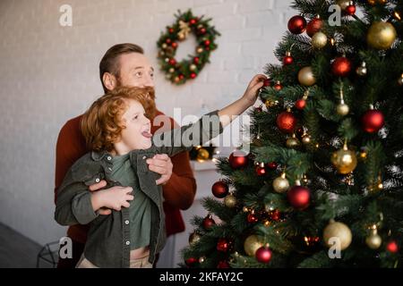 Mann hält Rotschopf Sohn aus der Zunge, während Dekoration Kiefer mit Weihnachtskugeln Stockfoto