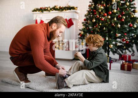 Seitenansicht des Rotkopfkindes, der auf dem Boden neben dem Vater sitzt, der seine Schnürsenkel und seinen Weihnachtsbaum mit einem dekorierten Kamin bindet Stockfoto