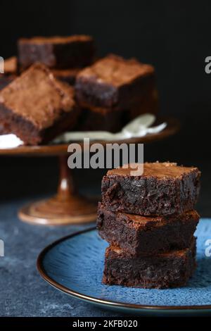 Ein Stapel hausgemachter, köstlicher Schokoladenbrownies auf einem goldverzierten blauen Teller vor dunklem Hintergrund mit mehr Brownies auf einem klassischen Dunkel Stockfoto