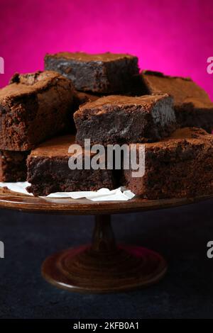 Ein Haufen hausgemachter, leckerer Schokoladenbrownies auf einem alten Holzkuchenstand vor einem dunkelrosa Hintergrund, ideal für den Valentinstag Stockfoto