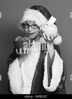 Junge süße weihnachtsmann Junge mit Gläsern in Weihnachten Pullover und Neujahr weihnachtshut mit rosa Schweinebank und Schokolade Hip Cookies in Schüssel auf Stockfoto