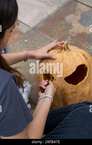 Frau schnitzt großen orangefarbenen Kürbis für Halloween, während sie zu Hause am Holztisch sitzt Stockfoto