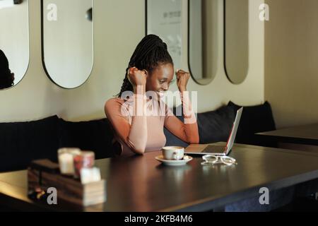 Junge afroamerikanische Frau, die im Café in der Nähe des Laptops am Tisch sitzt, die Fäuste hebt, Erfolge ausdrückt, gewinnt, feiert. Stockfoto