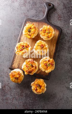 Frisch gebackene Blätterteigbecher gefüllt mit Eiern, Käse und Schinken aus nächster Nähe auf einem Holzbrett auf dem Tisch. Vertikale Draufsicht von oben Stockfoto