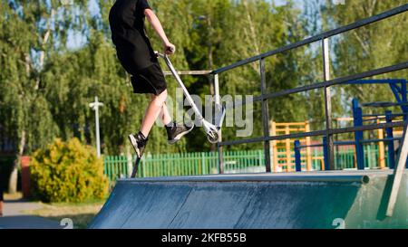 Ein Teenager in einem schwarzen T-Shirt und Shorts, die an einem sonnigen Tag auf einem Roller in einem Skatepark laufen Stockfoto