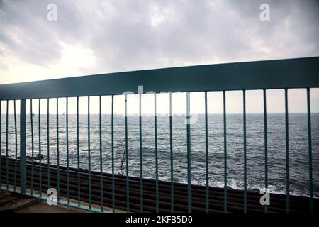 Meer mit Segelbooten in der Ferne an einem bewölkten Tag durch ein blaues Geländer gesehen Stockfoto