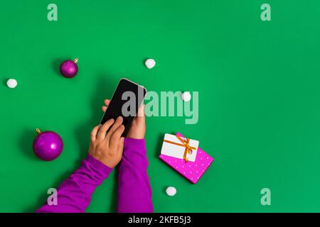 Hände in einem violetten Pullover berühren den Smartphone-Bildschirm mit den Fingern, Online-Zahlung für Weihnachtseinkäufe, grüner Hintergrund, flach liegend. Heller Koncep Stockfoto