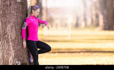 Eine junge Sportlerin mit einer athletischen Figur lehnt sich im Park an einen Baum und überprüft beim Laufen ihre Herzfrequenz und hört dabei auch Musik Stockfoto