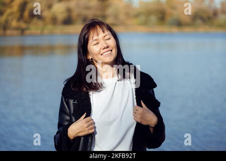Porträt einer Frau mittleren Alters in schwarzer Jacke, weißem T-Shirt, stehend mit geschlossenen Augen in der Nähe des Flusses, das Wetter genießen. Stockfoto