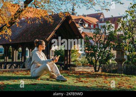 Junge Modische Teenage-Mädchen Mit Smartphone Im Europäischen Park Im Herbst Sitzen Lächelnd. Trendige Junge Frau Im Herbst In Park Texting. Retuschiert Stockfoto