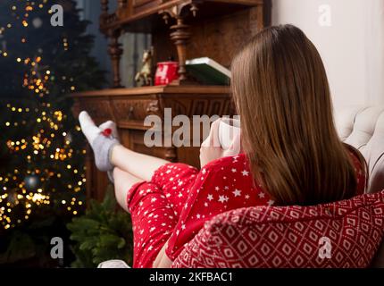 Weibliche Beine mit Füßen in niedlichen Weihnachtssocken, die sich bis zum Weihnachtsbaum erstrecken, im gemütlichen, warmen Zuhause. Hygge-Konzept. Stockfoto