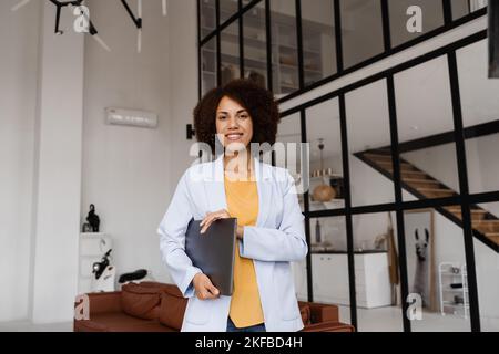 Selbstbewusste afrikanische Frau makler mit Laptop zeigt Wohnungen zum Mieten oder Kaufen. Hypothek für Wohnung für Kunden. Kreatives Foto für das Immobilienalter Stockfoto