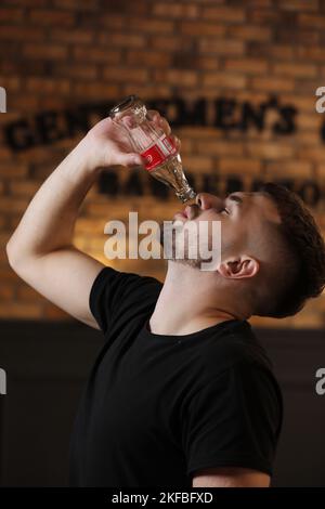 RIVNE, UKRAINE JUNI 26 2020: Junger bärtiger Mann trinkt Coca Cola aus der Flasche im Friseurladen auf Backsteinmauer Hintergrund. Stockfoto