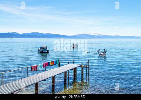 Dock mit Strandhandtüchern, die sich auf den Tahoe See ausdehnen, mit Booten, die vor der Küste festgemacht sind, und schneebedeckten Bergen und einem pastellfarbenen Himmel in der Ferne - B Stockfoto