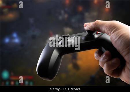 Ein Spieler hält vor dem Hintergrund eines Videospiels auf einem großen Plasma-TV-Bildschirm ein modernes GAM22ICB Gamepad in der Hand. Online-Spiele mit Freunden, com Stockfoto