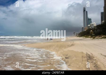 Reifenspuren und nicht identifizierbare Touristen am Strand, die am stürmischen Tag mit dunklen Wolken an der Gold Coast - Surfers Paradise Australia mit C ins Meer watten Stockfoto