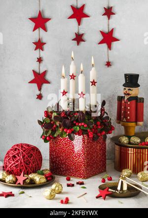 Stimmungsvolle Blumendekoration zu Weihnachten. Rote Waldbeerpflanze mit vier weißen brennenden Kerzen und Holzsternen in einem Blechtopf. Stockfoto