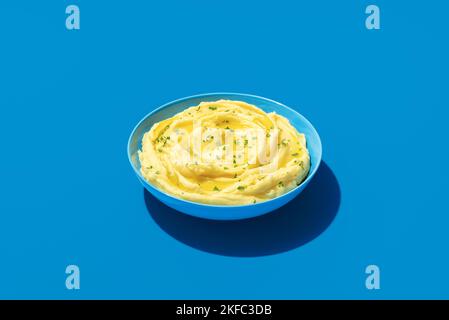 Cremige Kartoffelpüree mit grüner Petersilie in hellem Licht auf einem blauen Tisch. Schüssel mit Kartoffelpüree minimalistisch auf einem farbenfrohen Hintergrund Stockfoto