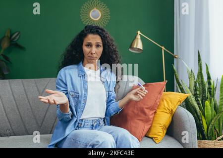 Eine junge, schöne lateinamerikanische Frau erhielt einen negativen Schwangerschaftstest. Sie sitzt aufgeregt zu Hause auf der Couch, blickt auf die Kamera, spreizt ihre Hände. Stockfoto