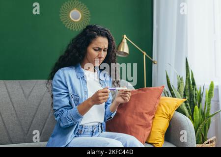 Eine junge, schöne lateinamerikanische Frau erhielt einen negativen Schwangerschaftstest. Sie sitzt aufgeregt zu Hause auf der Couch und hält einen Test in ihren Händen. Stockfoto