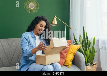 Eine junge lateinamerikanische Frau erhielt ein angenehmes Paket, das per Kurier zu ihrem Haus geliefert wurde. Sie sitzt glücklich auf dem Sofa und packt die Schachtel aus. Stockfoto