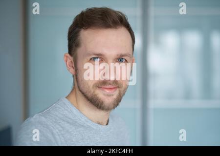 Nahaufnahme eines jungen hübschen kaukasischen Mannes in grauem T-Shirt, der in die Kamera schaut, positive Emotionen - lächelnd. Attraktiver bärtiger Mann im Heimbüro. Smart-Tech-Konzept. Hochwertige Bilder Stockfoto
