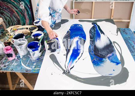 Beschnittenes Foto von Händen einer Frau in einem Gewand, bedeckt mit Flecken, die Farbe aus dem Eimer auf Leinwand gießen, mit abstraktem Bild. Stockfoto