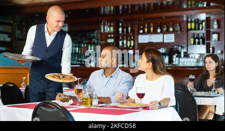 Freundlicher Kellner, der bestellte Pizzas zu Pärchen in der Pizzeria brachte Stockfoto