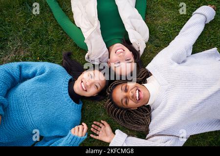 Fröhliches Porträt von drei besten Freunden, die auf dem Gras liegen und Spaß am Lachen haben und die Kamera betrachten Stockfoto