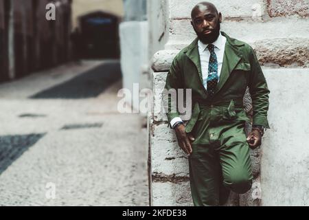 Ein schicker, tougher, kahlbärtiger, hübscher Schwarzer in einem modischen, grünlichen Outfit, der sich im Freien an die Wand lehnt; ein Straßenporträt eines afrikanischen Mannes Stockfoto
