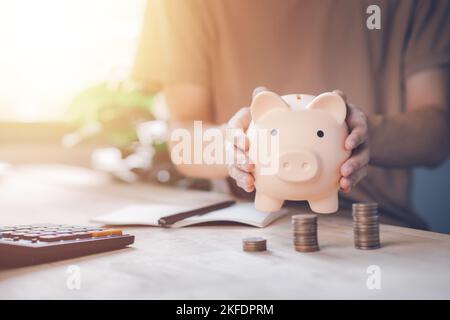 Mann Hand hält Sparschwein mit Rechner und Münze auf Holztisch. Geld sparen Reichtum und finanzielle Konzept. Unternehmen, Finanzen, Investitionen, Finanzen Stockfoto