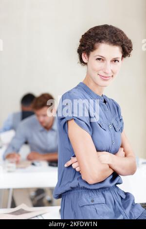 Wir nehmen Ihre IT-Bedürfnisse ernst. Porträt einer selbstbewussten Büroangestellerin, die auf ihrem Schreibtisch sitzt, mit Kollegen, die hinter ihr sitzen. Stockfoto