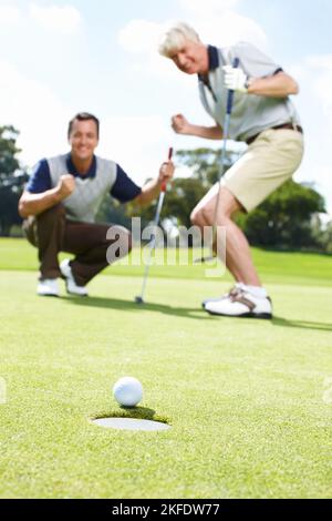 Ja. Zwei aufgeregt Männer auf dem Grün während einer Runde Golf beobachten den Ball ins Loch Rollen. Stockfoto