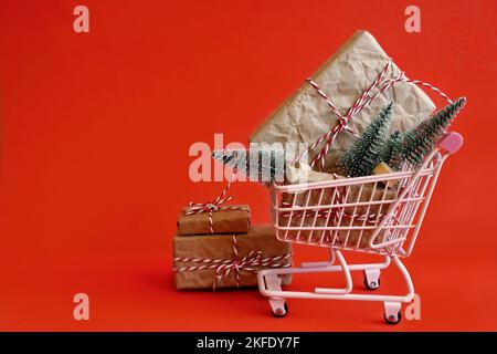 Weihnachtsangebot – Einkaufswagen und Weihnachtsgeschenke auf rotem Hintergrund. Frohe Weihnachten, frohes neues Jahr, Einkaufskonzept. Draufsicht und Kopierbereich. Stockfoto