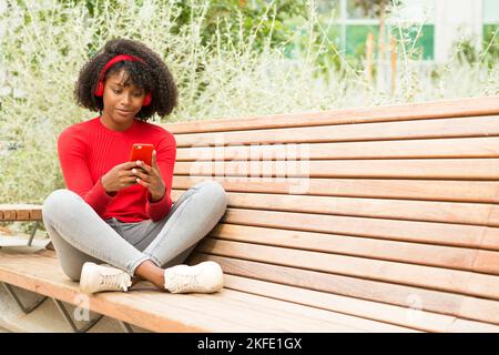 Junge afroamerikanische Frau, die mit Kopfhörern Musik hört, während sie das Telefon im Freien benutzt Stockfoto