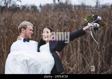 Lustige gerade verheiratet heterosexuellen Paar. Starker skandinavischer Mann, der seine lachende türkische Braut hochhebt. Feld im Hintergrund. Herbsthochzeit. Hochwertige Fotos Stockfoto