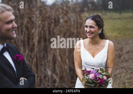 Lachende türkische Braut im ärmellosen Brautkleid, die während des Herbstfotoshootings im Freien über ihren Mann lachte. Beste Freunde als Ehepartner. Hochwertige Fotos Stockfoto