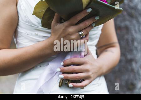 Nicht erkennbare türkische Braut im ärmellosen Brautkleid, die ihren Brautstrauß mit beiden Händen hält und ihren neuen Ehering und wunderschöne Nägel zeigt. Hochwertige Fotos Stockfoto