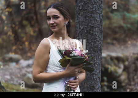 Mittelgroße Aufnahme einer attraktiven türkisch-braunen Braut im ärmellosen Brautkleid, die die Kamera anschaut und einen frischen Blumenstrauß hält. Verschwommener Hintergrund - Herbstwald. . Hochwertige Fotos Stockfoto
