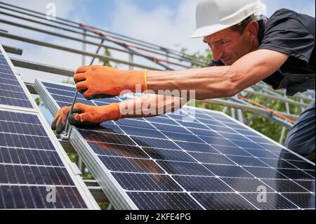 Nahaufnahme des man-Installateurs, der das Solarmodul auf Metallschienen platziert. Männlicher Arbeiter, der das Photovoltaik-Solarpanel-System im Freien montiert, trägt Bauhelm und Arbeitshandschuhe. Stockfoto