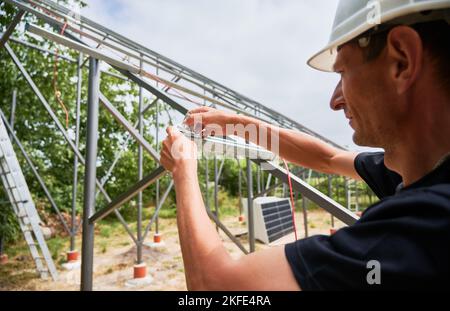 Arbeiter Anziehen Montageklammer in Metallträger für die Installation von Solarmodulen. Erneuerbare und ökologische Energie. Idee der Umwelt sicher. Moderne Technologie und Innovation. Mann mit Arbeitskleidung und Helm Stockfoto