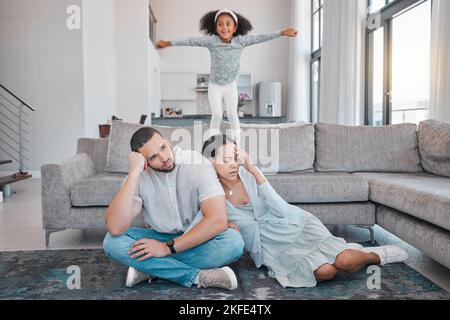 Müde springen Eltern und Kind auf das Sofa, während die erschöpfte Mutter und der erschöpfte Vater im Wohnzimmer auf dem Boden sitzen. Familie, Energie und junges Mädchen springen auf Couch Stockfoto